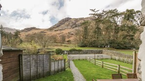 A patio with a table and chairs, a stone pathway, a wooden fence, and a view of a mountainous landscape. - Holly Tree Cottage (Coniston)