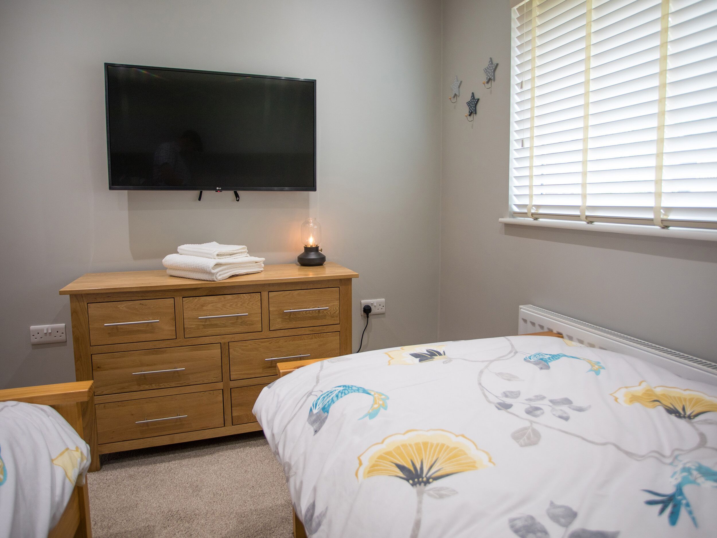 A bedroom with a wooden dresser, a flat-screen TV mounted on the wall, a bedside lamp, and a window with blinds.