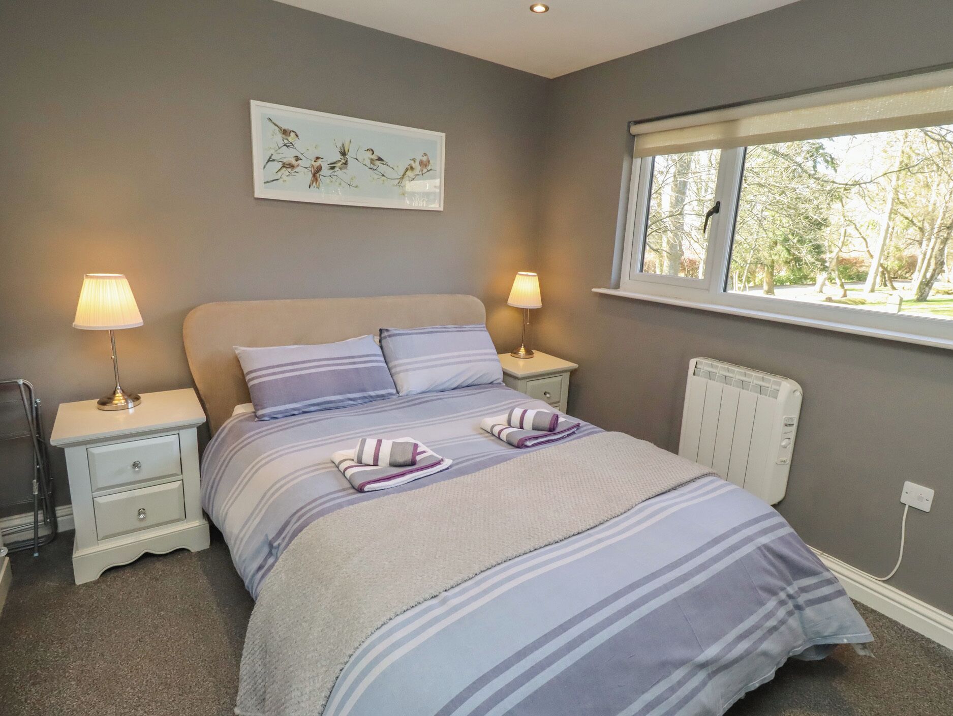 A bedroom with a bed, two bedside lamps, a radiator, a window with a view, and a wall-mounted artwork.
