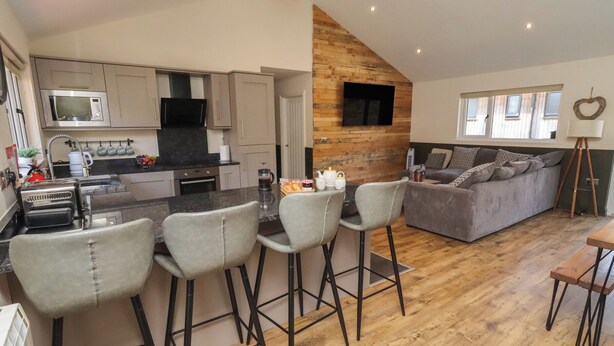 A modern kitchen with a central island, bar stools, a grey sofa, and a wooden accent wall. - Barn Owl (Newcastle-upon-Tyne)
