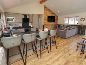 A modern kitchen with a central island, bar stools, a grey sofa, and a wooden accent wall. - Barn Owl (Newcastle-upon-Tyne)