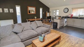 A modern living room with a sectional sofa, a wooden coffee table, a dining area with a table and chairs, a kitchen with a washing machine, and a large clock on the wall. - Barn Owl (Newcastle-upon-Tyne)