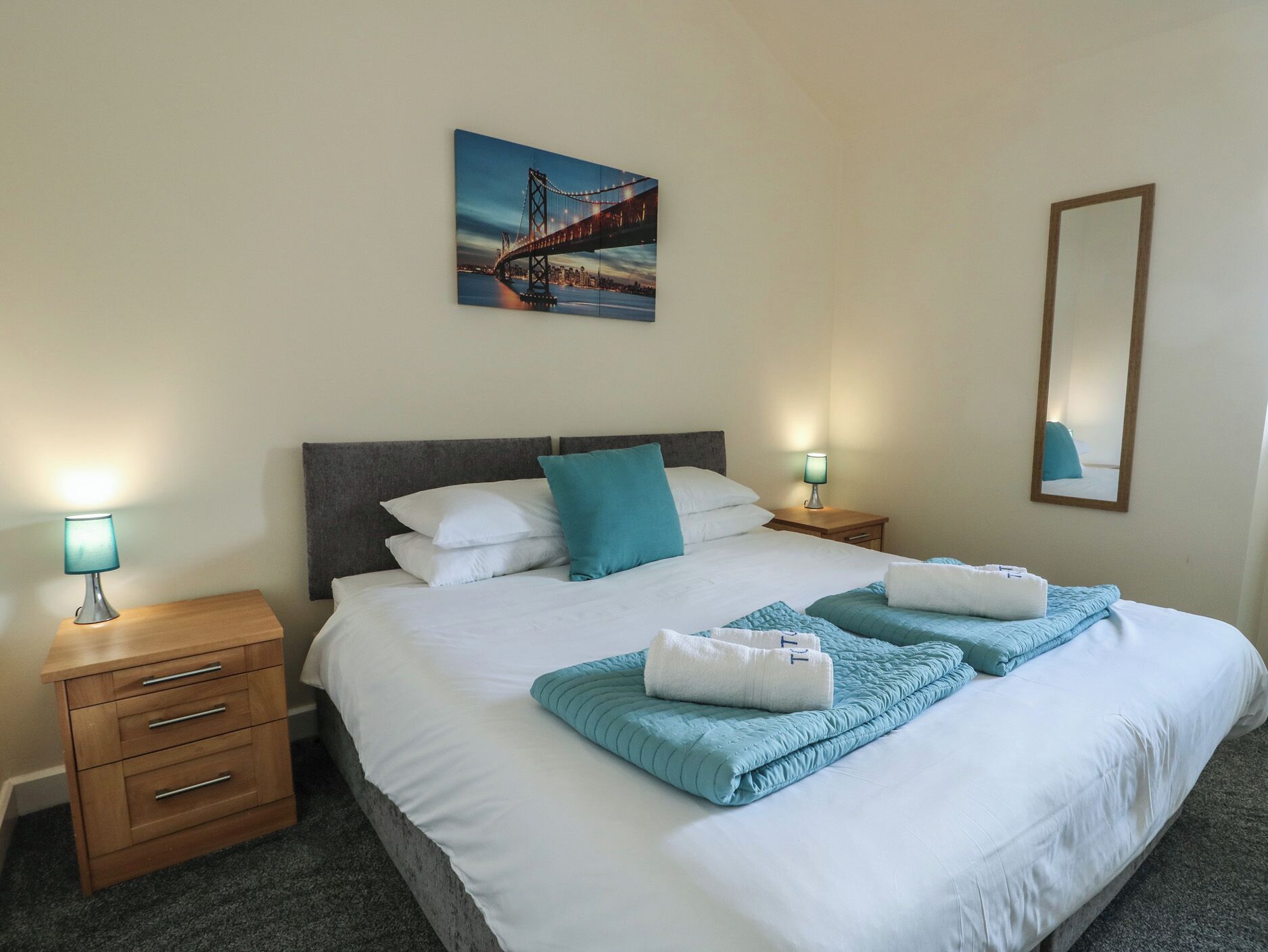 Uma cama bem arrumada com roupa de cama branca e azul-turquesa, uma mesa de cabeceira de madeira com uma luminária, um quadro de uma ponte e um espelho na parede.