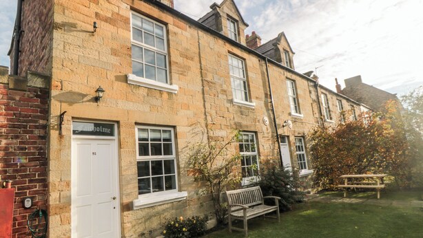 A traditional stone building with multiple windows, a front door, and a small garden area with a bench.
