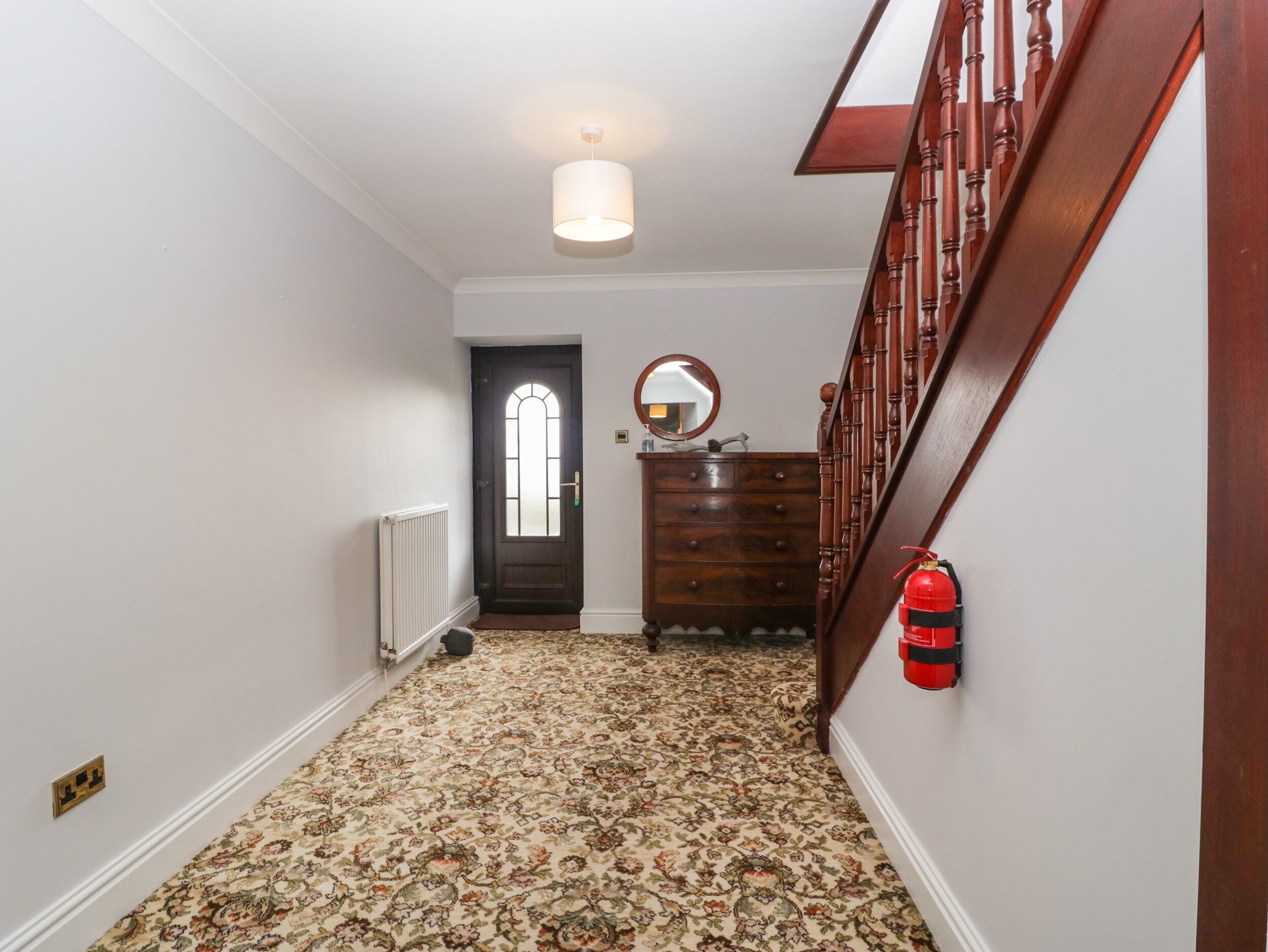 Un pasillo con alfombra estampada, una escalera de madera, una puerta oscura con ventanal, un aparador de madera y un detector de incendios rojo.