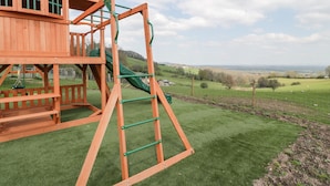 Una estructura de juego de madera con un tobogán y elementos para escalar, sobre un fondo de campo césped y colinas distantes.