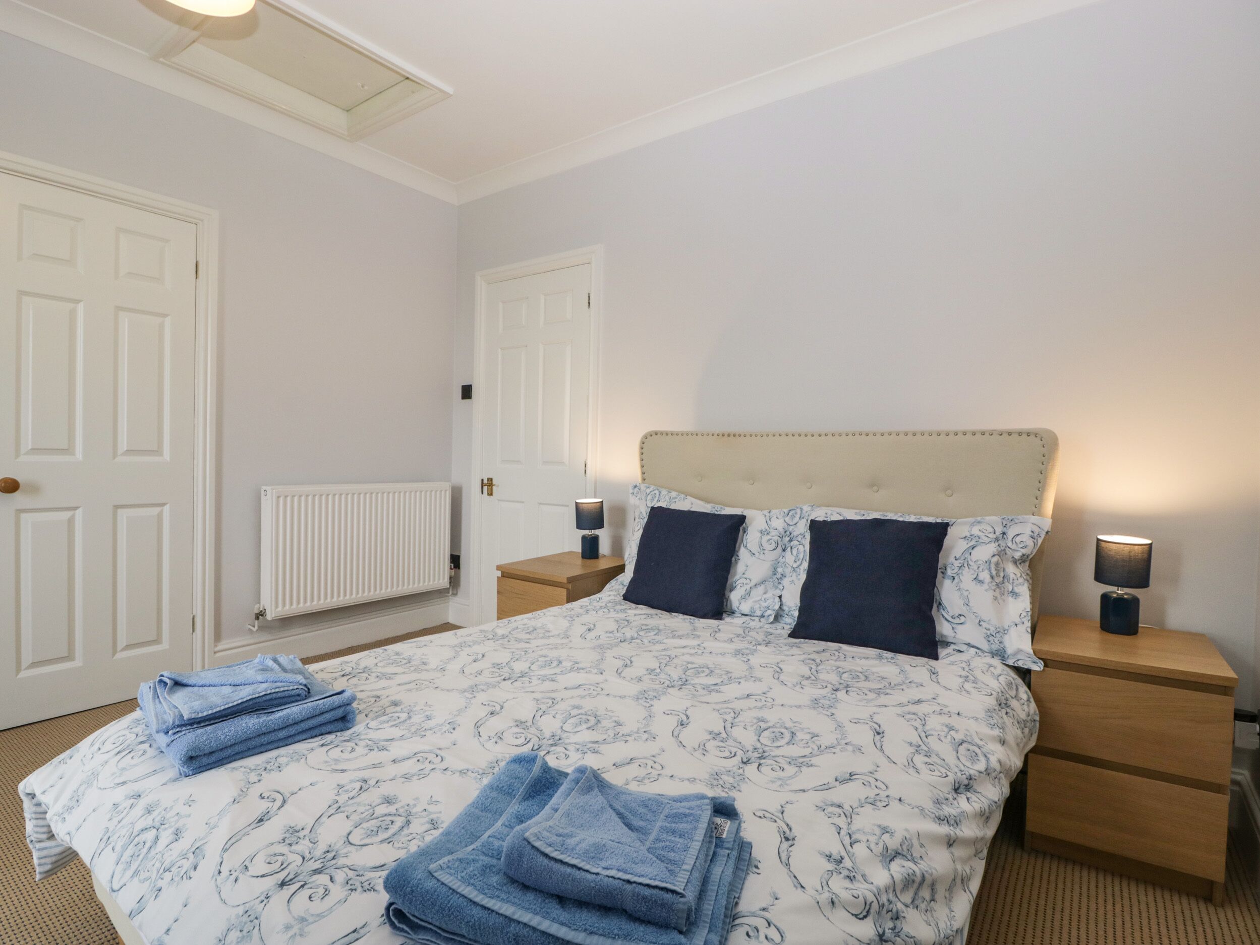 A neatly made bed with a patterned duvet, two bedside lamps, a wooden nightstand, and a radiator.