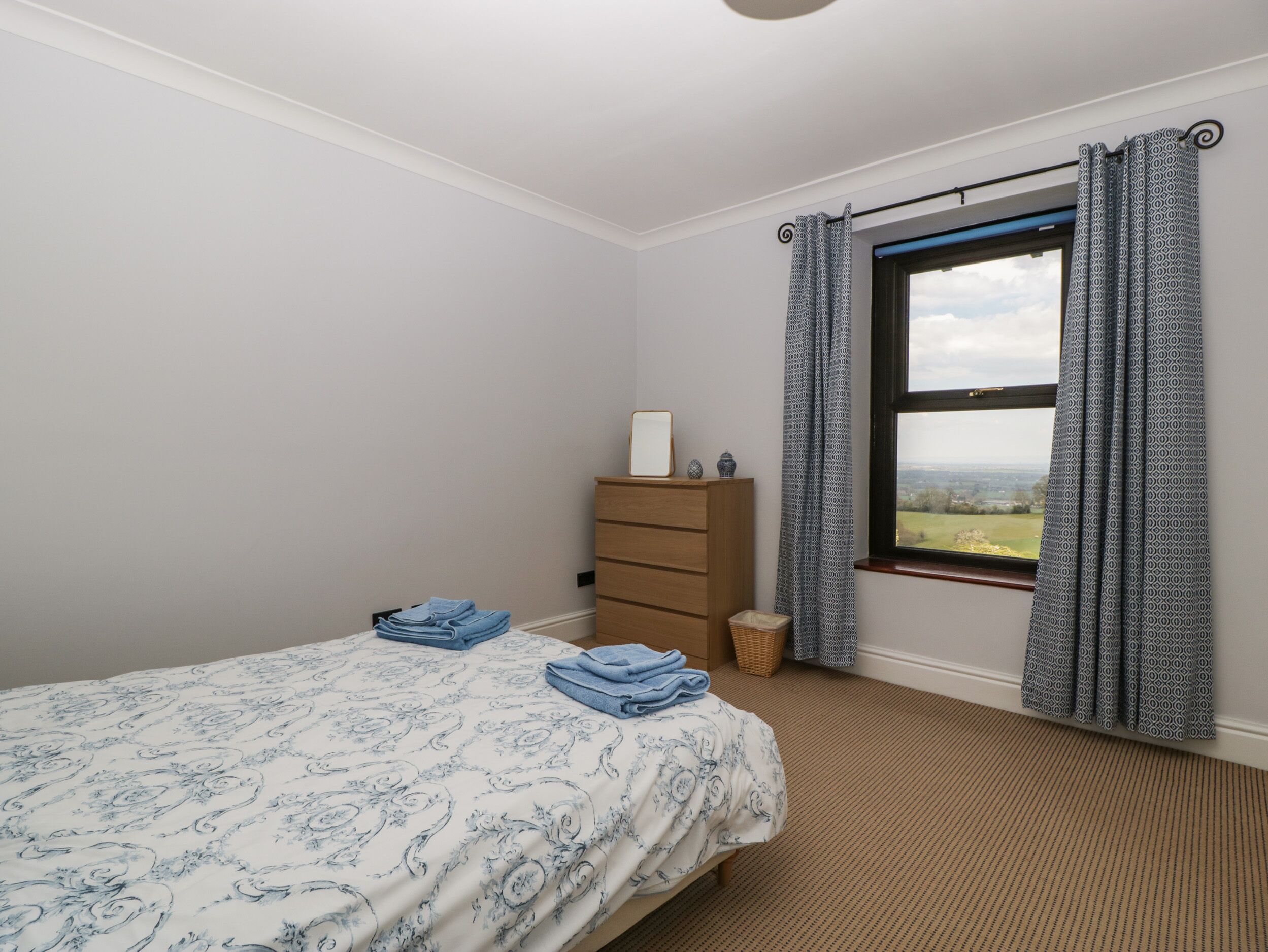 A bedroom with a bed, a dresser, a window with curtains, and a view of the countryside.