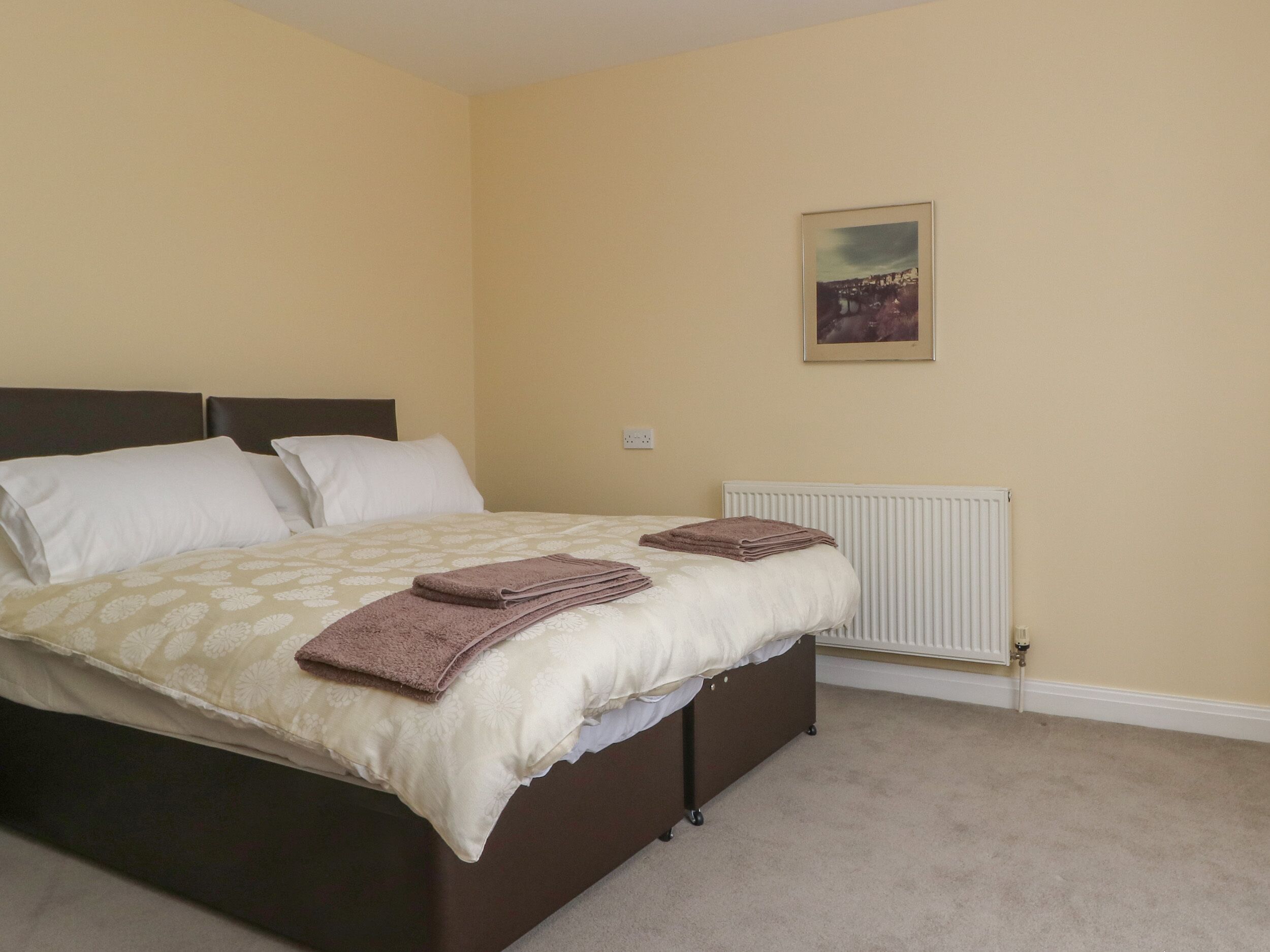 A neatly made bed with a patterned bedspread and two folded towels, a white radiator, and a framed picture on the wall.