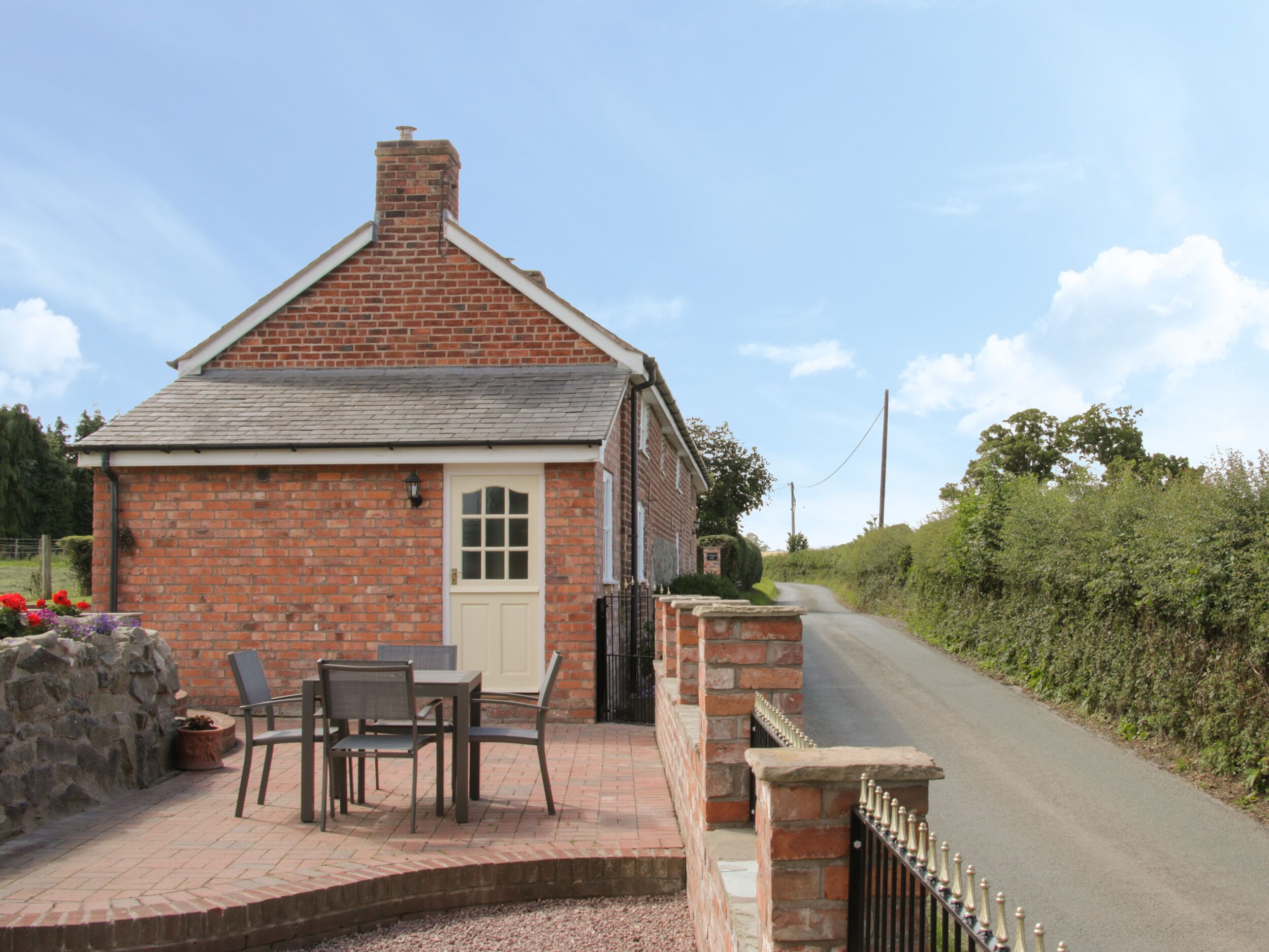 Une petite maison en briques avec une porte blanche et une cheminée. On y trouve une terrasse dallée avec une table et des chaises.