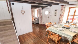 A dining area with a wooden table set for four, a staircase, and a living room with a sofa and floral curtains. - Pant y Ffynnon (Haverfordwest)