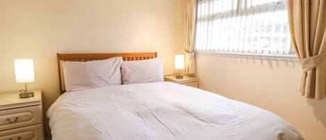 A neatly made bed with white linens, a wooden headboard, two bedside tables with lamps, and a window with curtains.