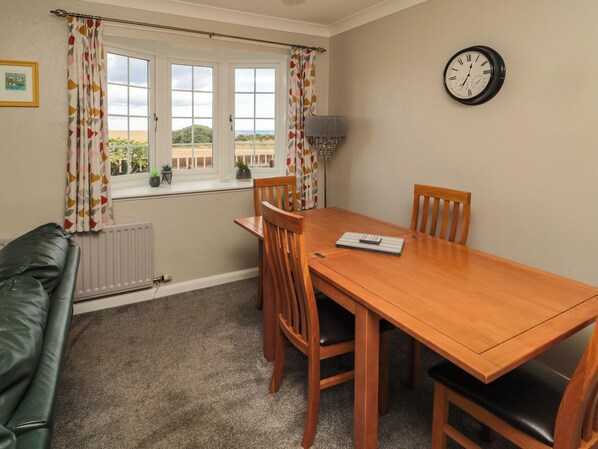 A dining area with a wooden table, four chairs, a clock on the wall, and a view through the window.