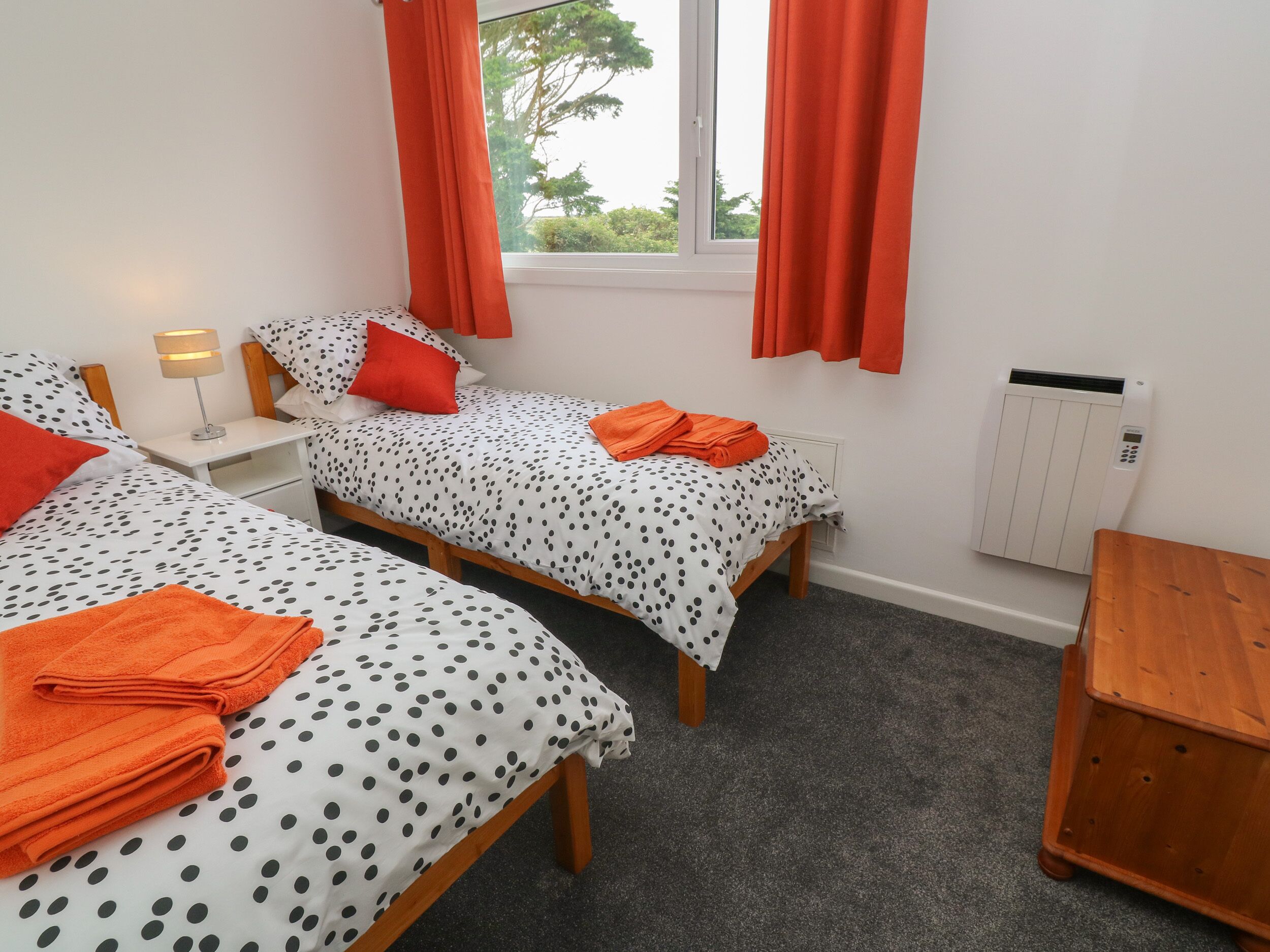 A bedroom with two beds, each with polka dot bedding and orange pillows. There is a window with orange curtains, a radiator, and a small table.