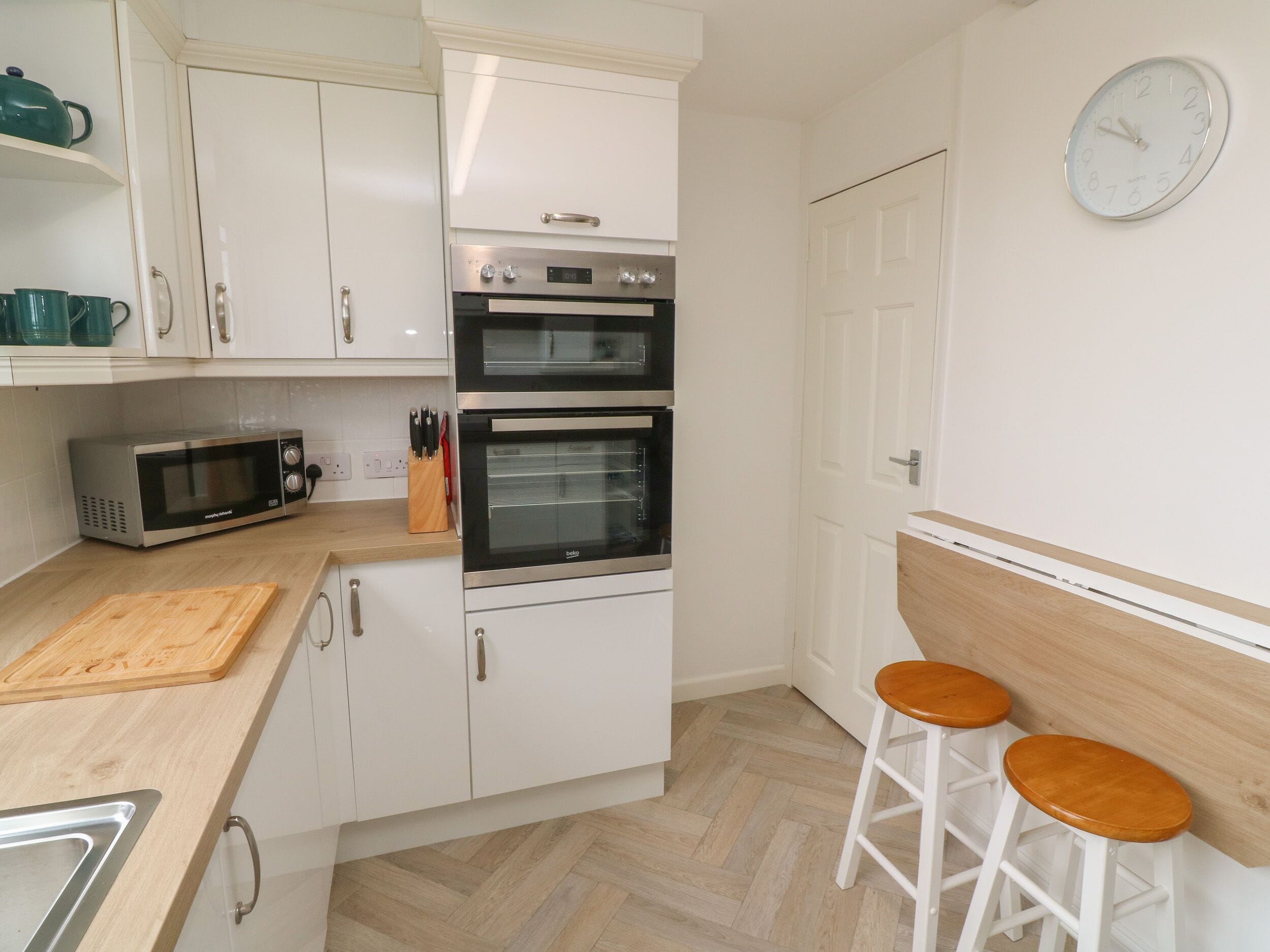 Een kleine keuken met witte kastjes, een magnetron, twee ovens, een houten snijplank en twee houten krukken.
