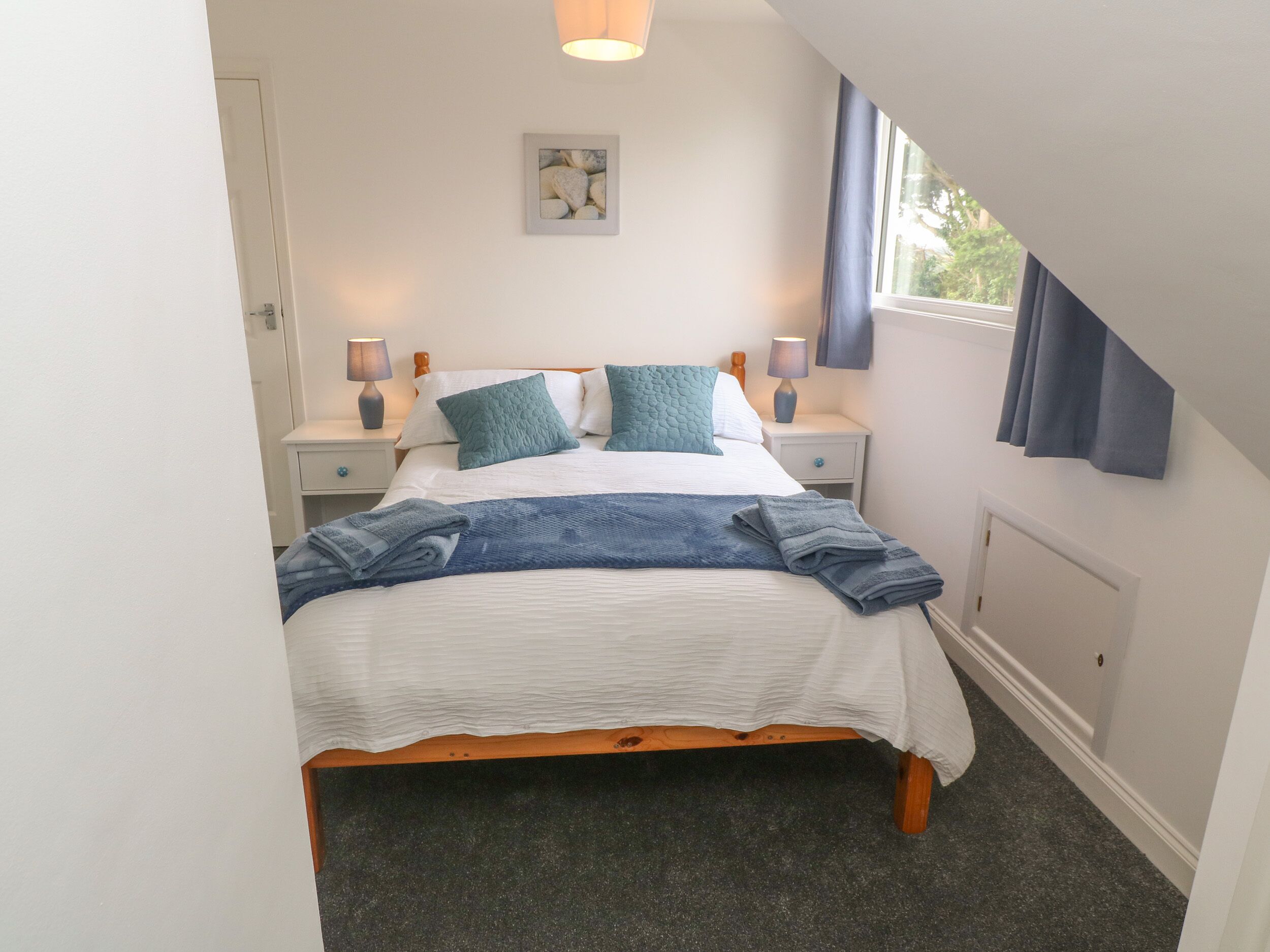 A neatly made bed with a white and blue duvet, two matching pillows, and a blue blanket. There are two bedside tables with lamps, and a window with curtains.