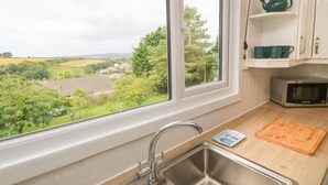 A kitchen with a stainless steel sink, a wooden cutting board, a microwave, and a window with a view of a green landscape. - Little Valley View (Hayle)