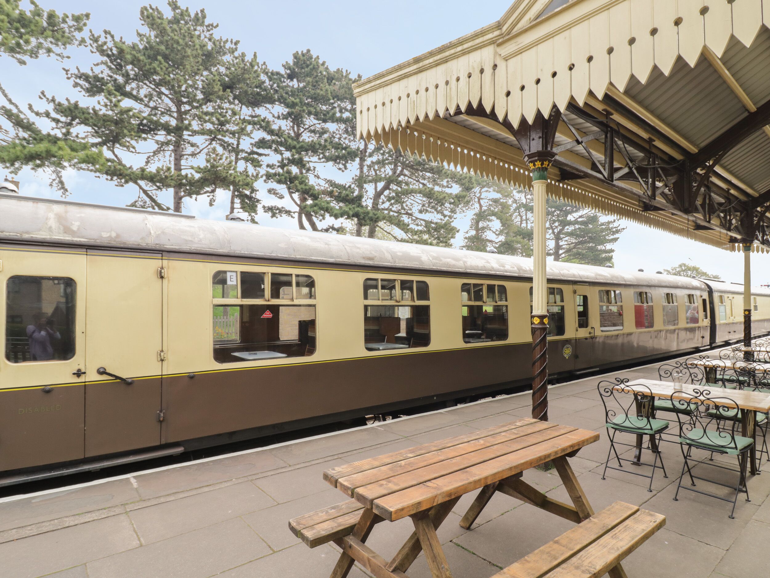 Un train d’époque dans une gare, avec des sièges à l’extérieur et des arbres en arrière-plan.