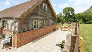 A wooden house with a sloped roof and brick foundation, a patio with outdoor furniture, and a fenced garden area.