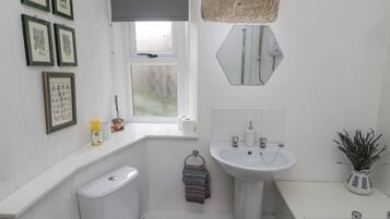 A bathroom with a toilet, sink, and bathtub. There are framed botanical prints on the wall, a window with a view, and a mirror above the sink.