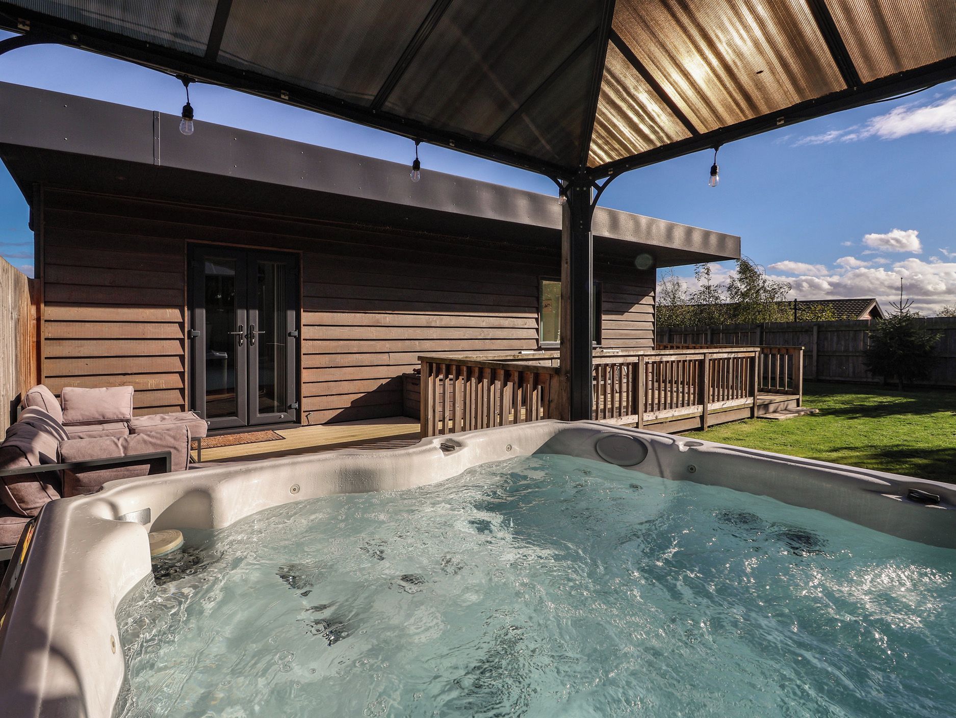 Un jacuzzi bajo un porche cubierto, con una casa de madera al fondo.