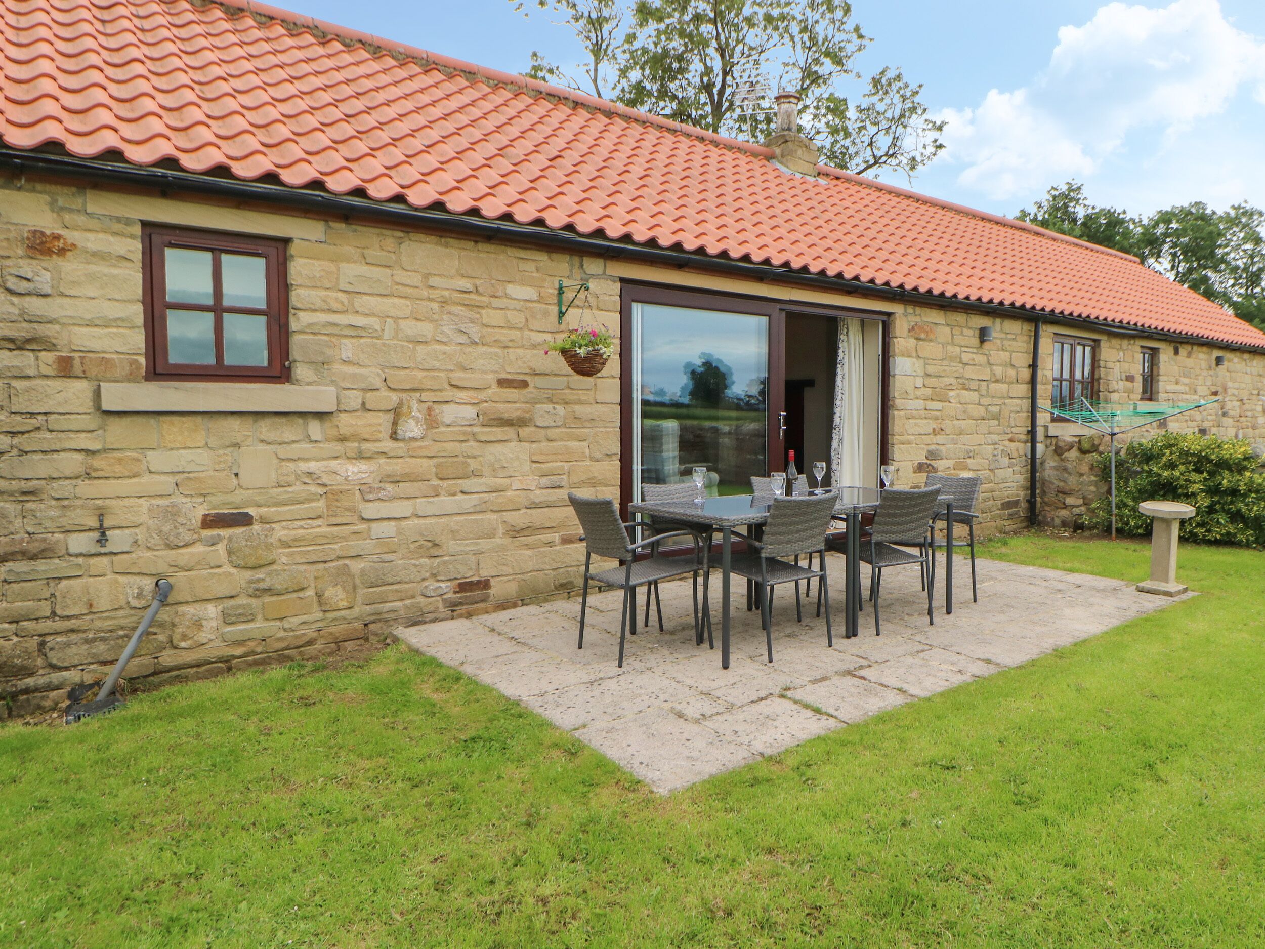 A stone building with a tiled roof, a patio with outdoor furniture, and a hanging flower basket.