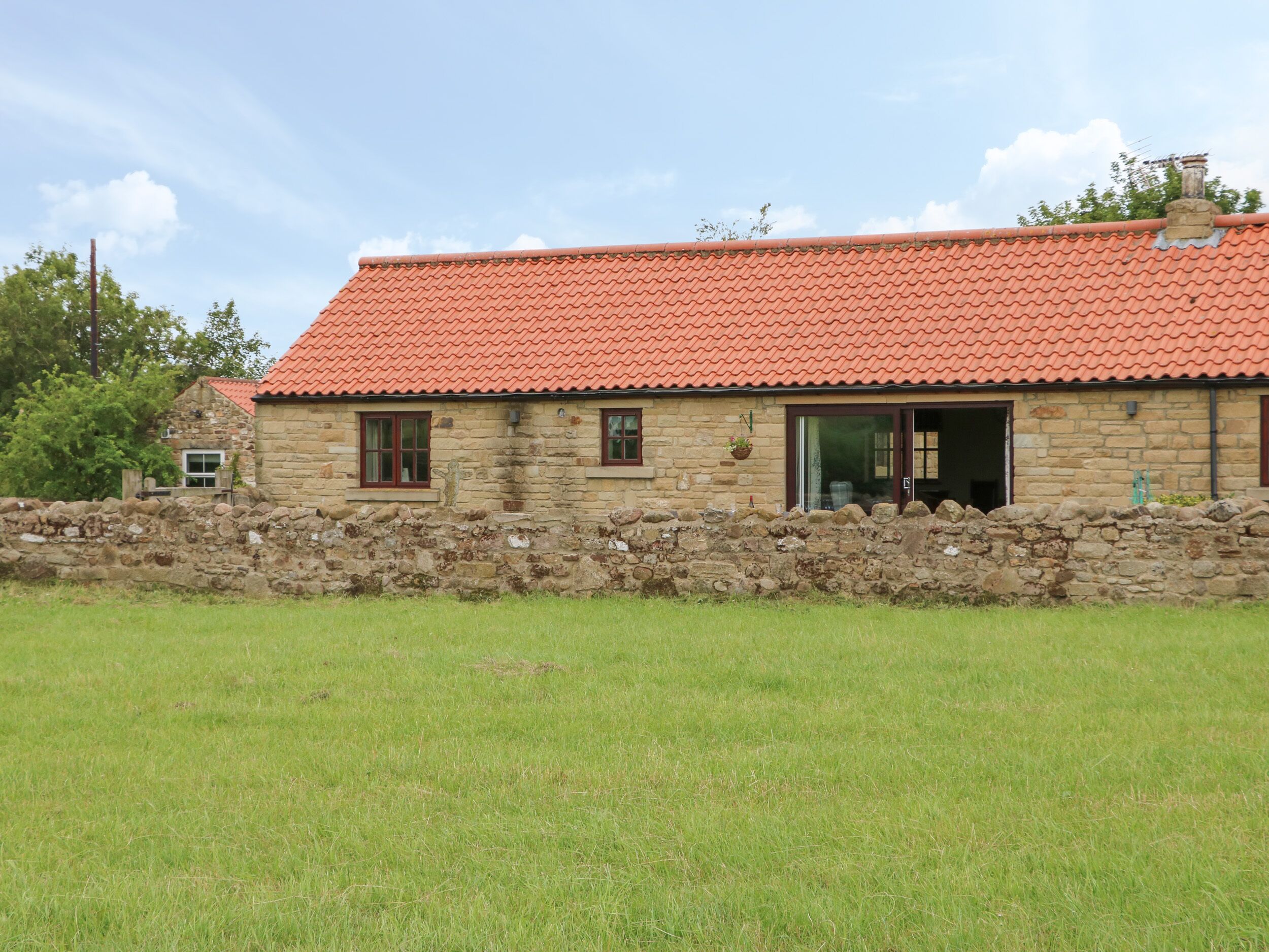 A stone building with a red-tiled roof, a stone wall, and a grassy field in the foreground.