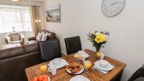Un comedor con una mesa de madera dispuesta para el desayuno, que incluye cruasanes, jugo de naranja y un bol de naranjas. Hay un reloj en la pared y un cuadro sobre la mesa.