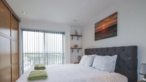 A bedroom with a large bed, a view of the outside through a window, and a painting on the wall. - Crantock Reach (Newquay)