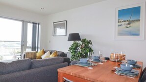 A modern living room with a grey sofa, a wooden dining table set for four, and a large window offering a view of the city. - Crantock Reach (Newquay)