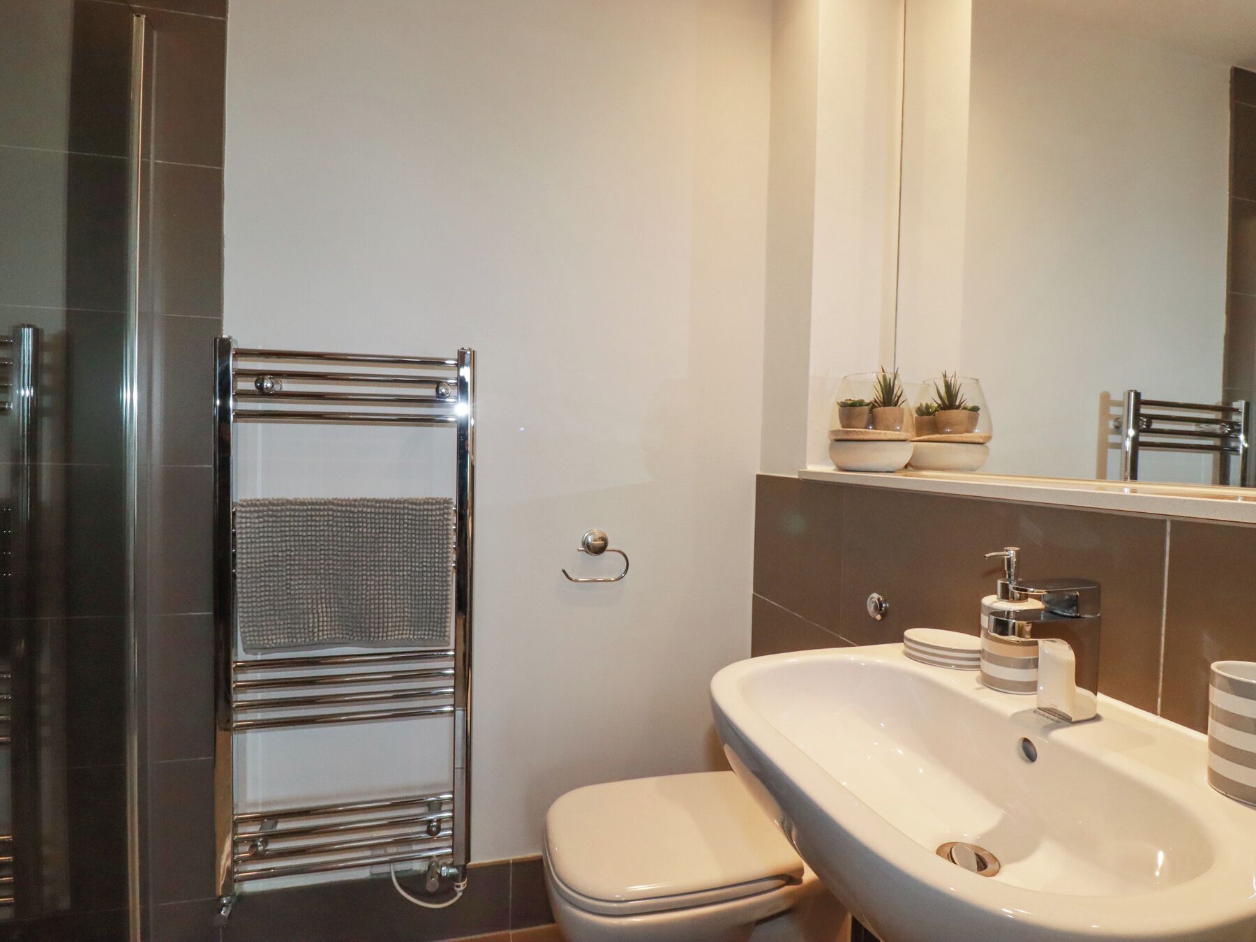Un baño moderno con un perchero de cromo, un inodoro blanco, un lavamanos grande con un espejo arriba y plantas decorativas en el mueble.