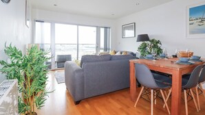 A modern living room with a grey sofa, wooden dining table, and chairs. There is a large window providing a view of the city. - Crantock Reach (Newquay)