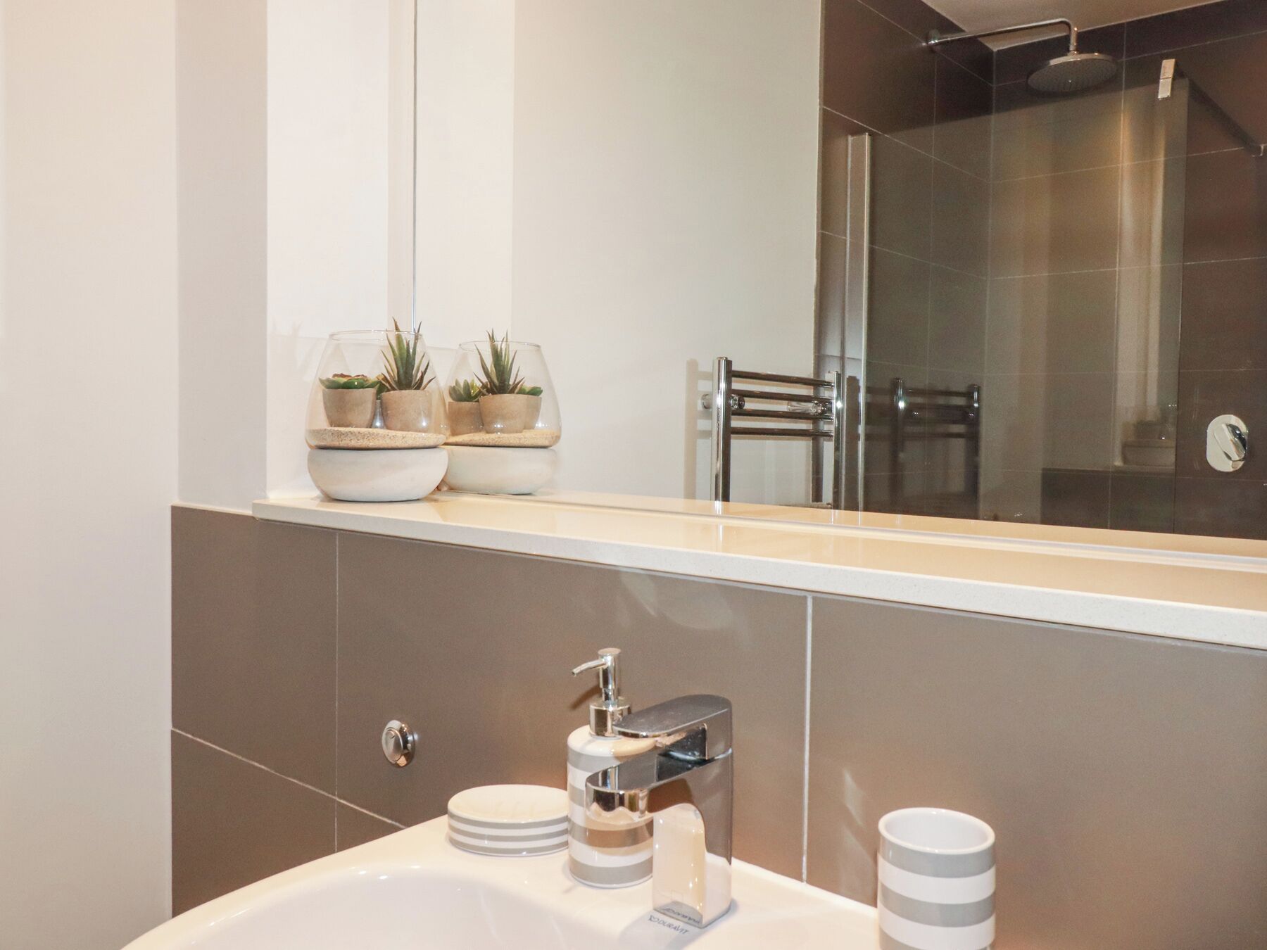 A modern bathroom with a large mirror, a sink with a chrome faucet, and two small potted plants on the countertop.