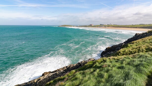 A coastal landscape with a rocky shoreline, grassy areas, and a distant building. - Crantock Reach (Newquay)