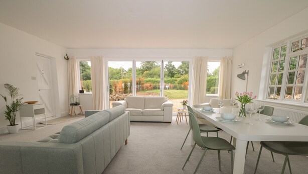 A modern living room with a grey sofa, a white dining table set with plates and glasses, and a view of a garden through large windows. - Thames Reach (Wallingford)