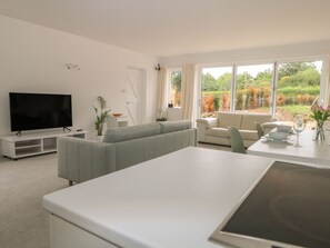 A modern living room with a flat-screen TV, a grey sofa, a dining table with chairs, and a large window with a view of trees. - Thames Reach (Wallingford)