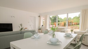 A modern living room with a dining table set for two, a grey sofa, and a large window offering a view of a garden. - Thames Reach (Wallingford)