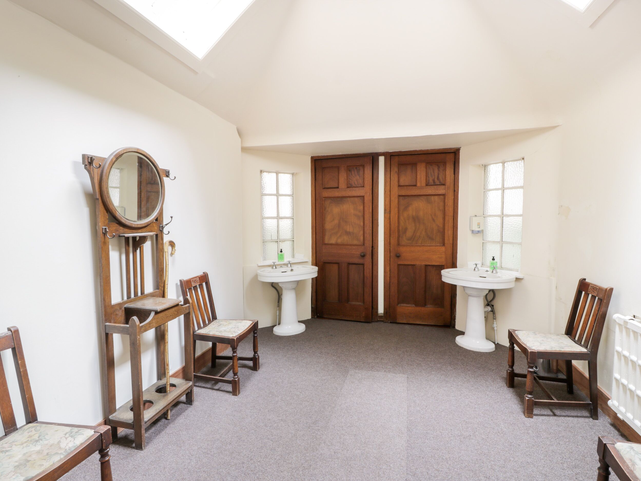 Una habitación con dos puertas de madera, un tragaluz, un espejo, dos lavamanos y cuatro sillas de madera.