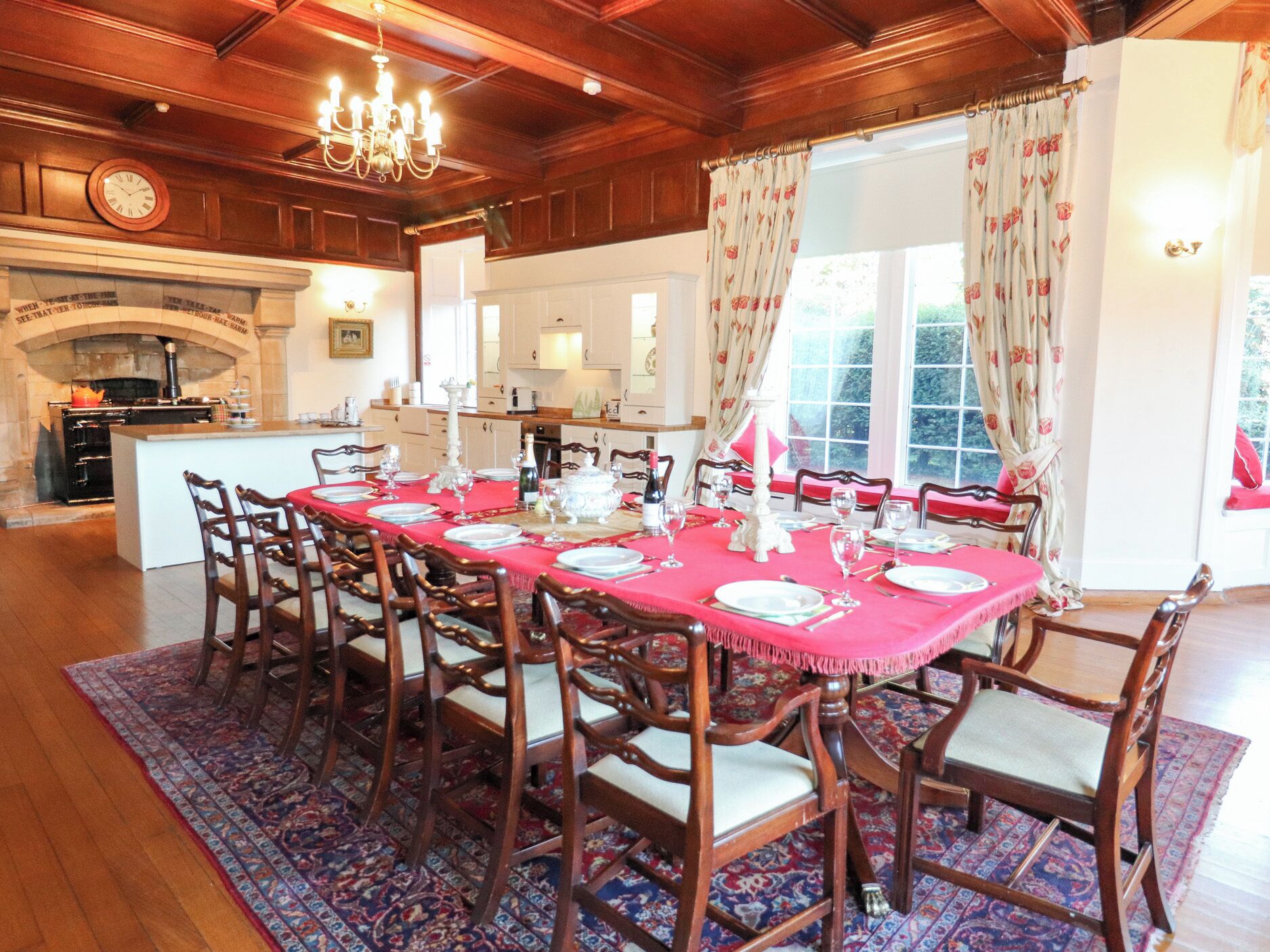 A dining room with a large table set for a meal, wooden chairs, a chandelier, and a kitchen in the background.
