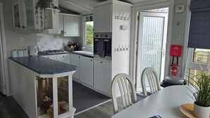 A modern kitchen with white cabinetry, a central island, and a dining area with a table and chairs. - Caban Tywod (Pwllheli)