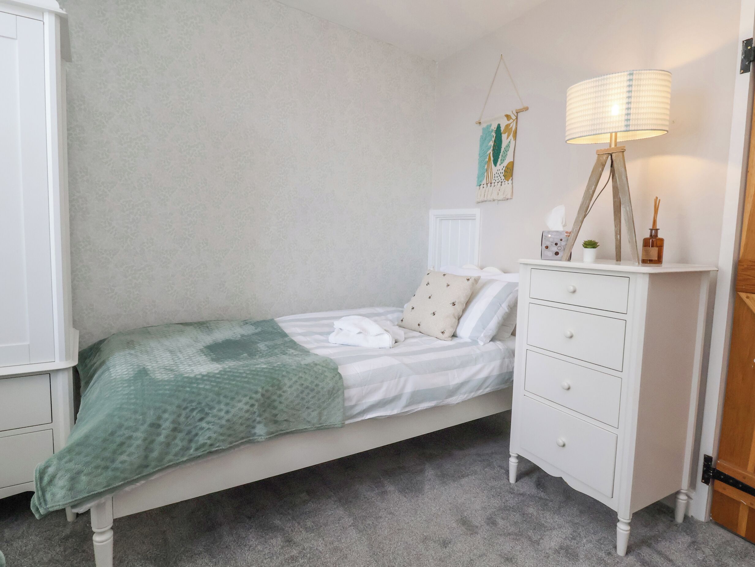 A bedroom with a single bed, a white dresser, a lamp, and a wall hanging.