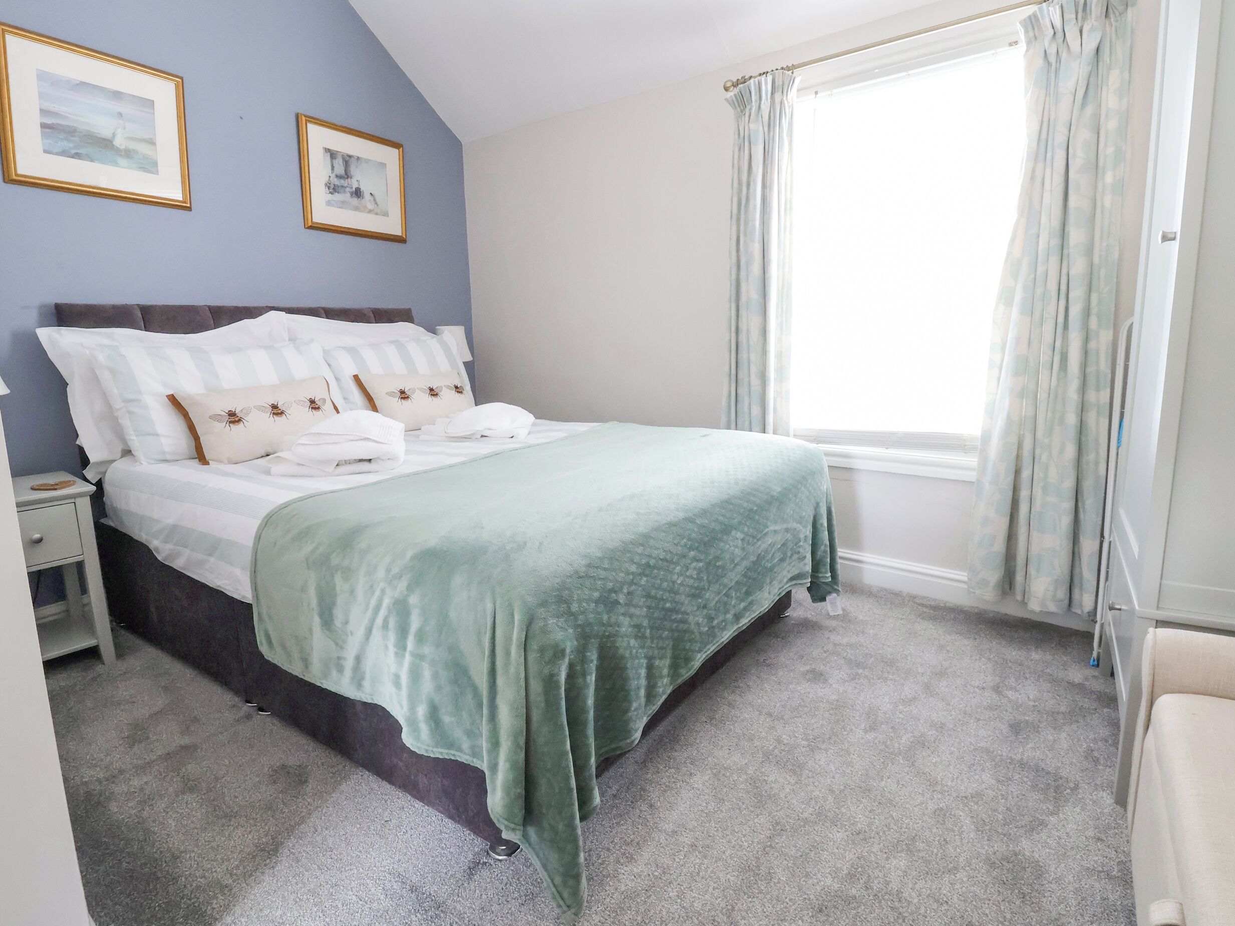 A neatly made bed with a green blanket, a nightstand with a lamp, and two framed pictures on the wall.