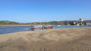 Escena costera con barcos amarrados en un muelle, una playa arenosa y un puente al fondo.