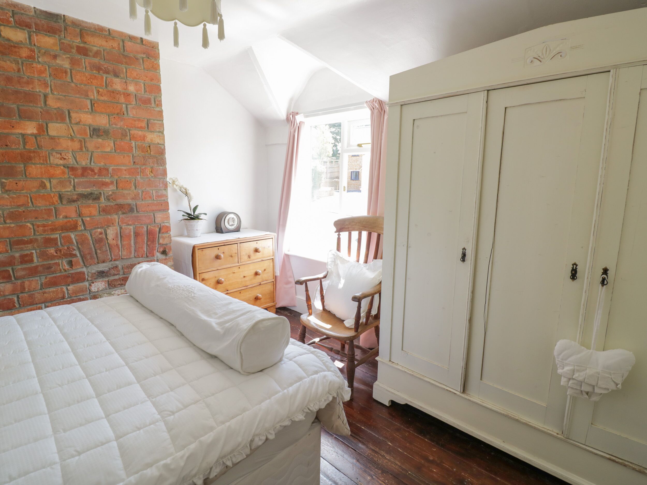 A bedroom with a brick wall, a wooden dresser, a chair, a wardrobe, and a bed with a quilted mattress.