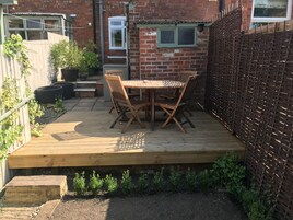 Una terraza de madera con una mesa y sillas, rodeada por un muro de ladrillo y una cerca trenzada.