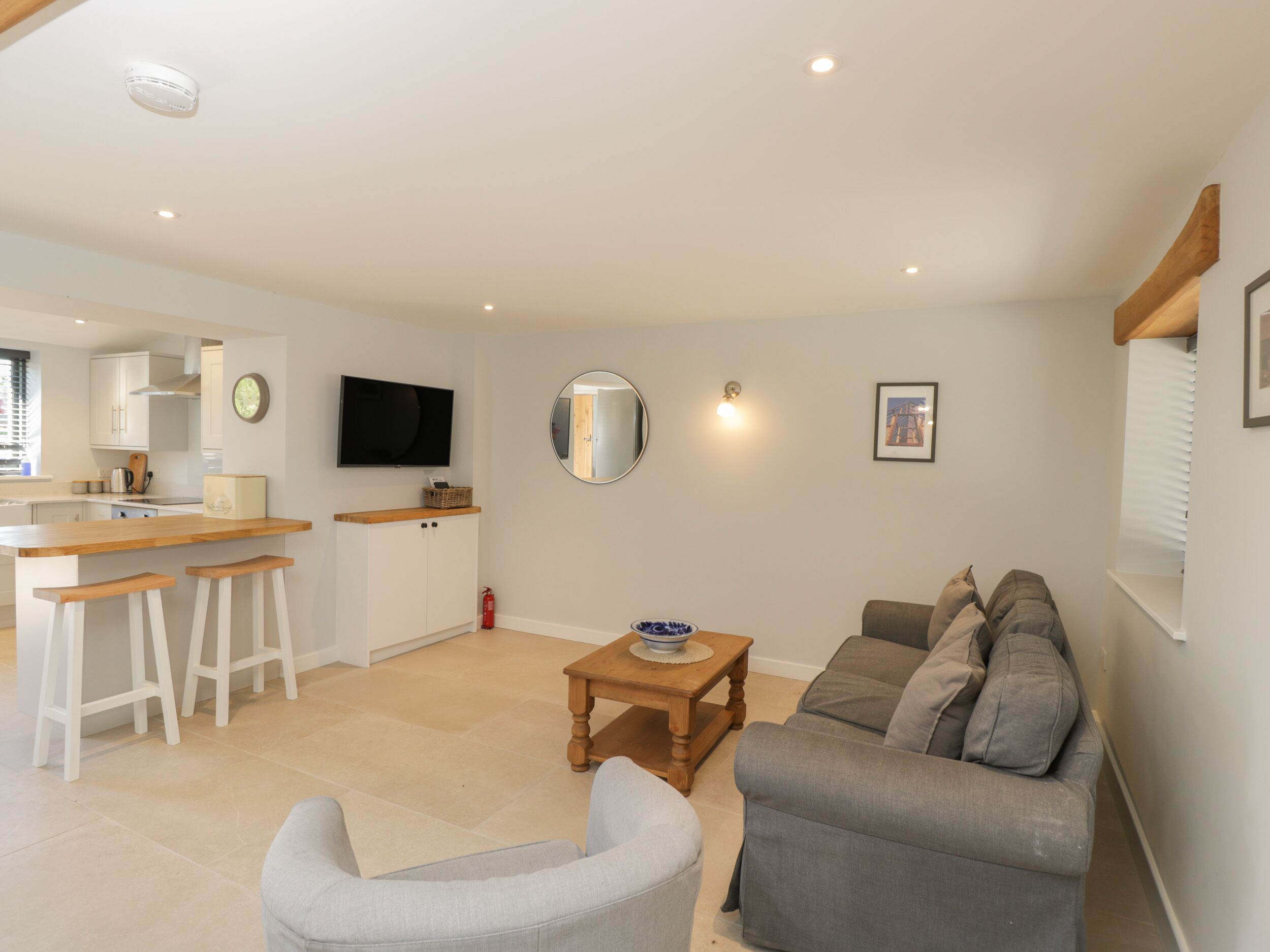 A modern living room with a grey sofa, a wooden coffee table, and a kitchen area with white cabinets and a flat-screen TV.