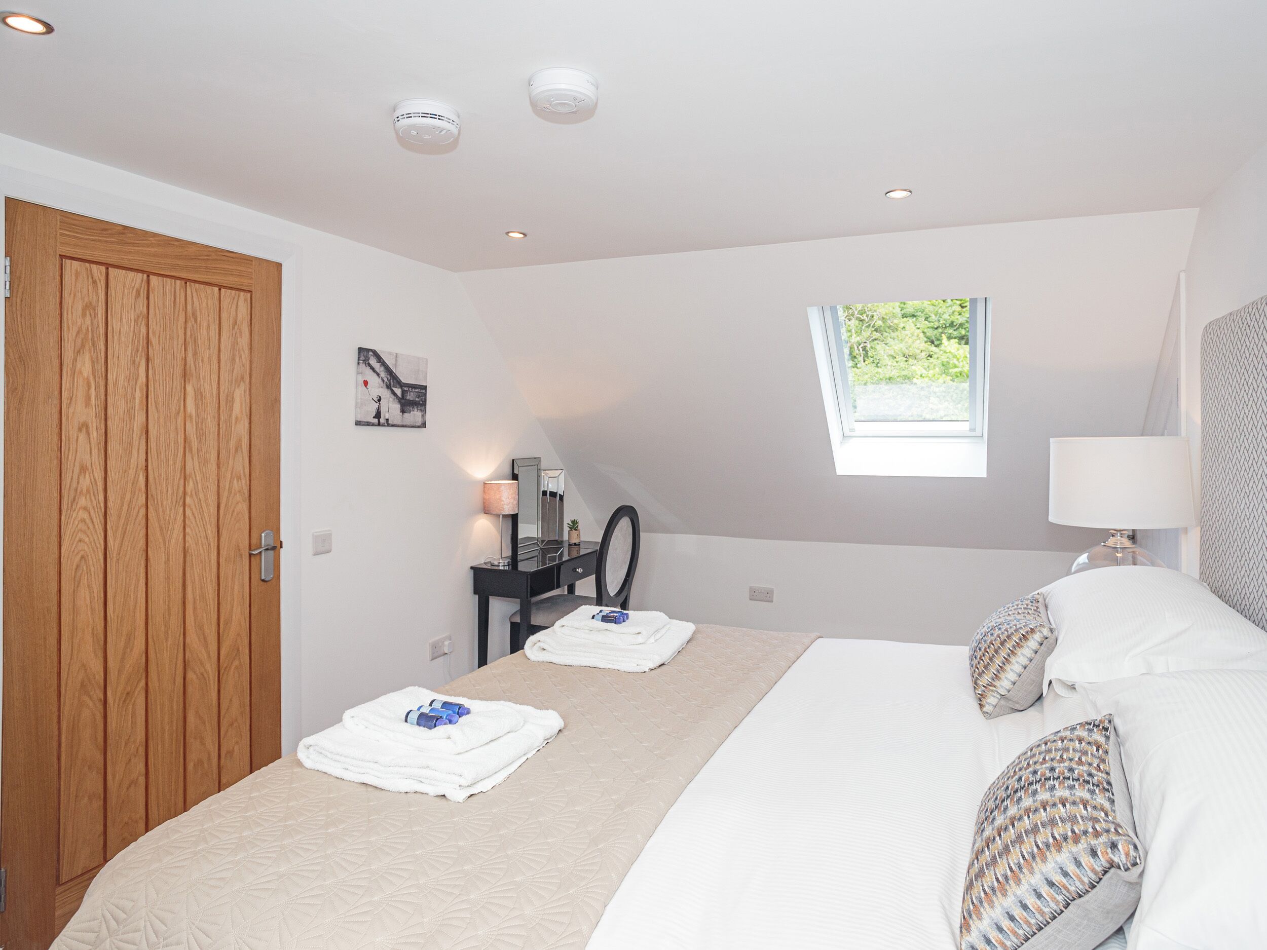 A bedroom with a bed, a wooden door, a small desk with a mirror, a window, and a ceiling light.