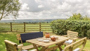 Une table de pique-nique en bois avec des bancs, un gâteau, une théière et des tasses, installée dans un champ verdoyant avec vue sur des collines.