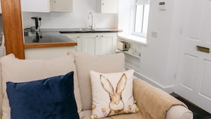Un salon chaleureux avec un canapé beige, un coussin bleu foncé et un coussin à motifs de lapin. On aperçoit, au fond, une cuisine avec des armoires blanches et un comptoir noir.