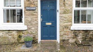 Un bâtiment en pierre avec une porte bleue, une plaque portant l’inscription « Pine Cottage », et un pot de fleurs placé devant.